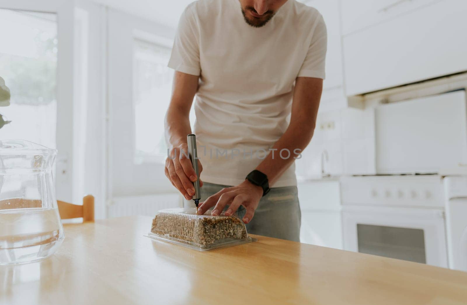 One young Caucasian recognizable man drills a plastic transparent box with filler for planting wheat with a mini-drill, standing in the kitchen at a wooden table, close-up view from below.