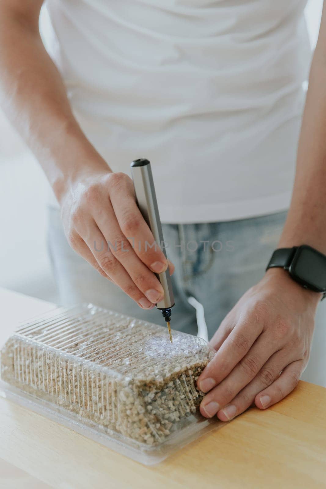 One young Caucasian unrecognizable man drills a plastic transparent box with filler for planting wheat with a mini-drill, standing in the kitchen at a wooden table, close-up view from below.