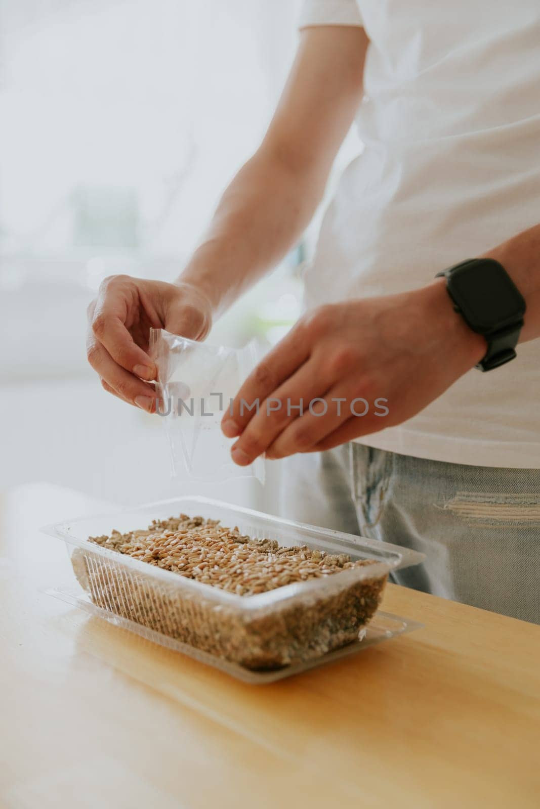 One young Caucasian unrecognizable man pours seeds from a transparent bag into a container with soil with both hands, standing in the kitchen at a wooden table, close-up side view.