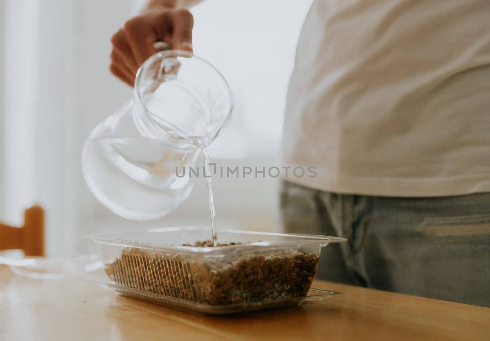 One young Caucasian unrecognizable man waters wheat seeds with soil into a container with one hand from a glass jug, standing in the kitchen at a wooden table, close-up side view.