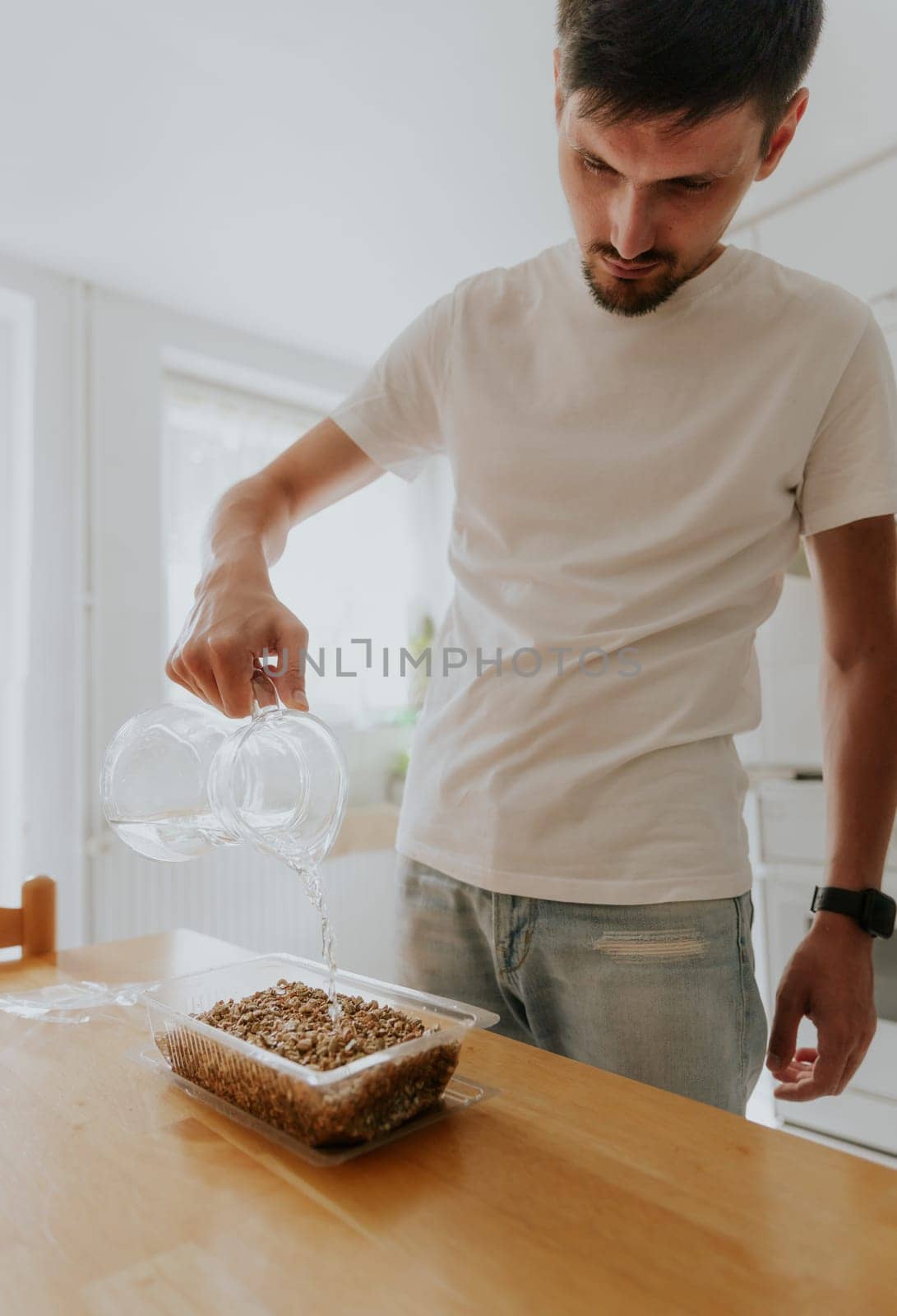 One young Caucasian recognizable man waters wheat seeds with soil into a container with one hand from a glass jug, standing in the kitchen at a wooden table, close-up side view.
