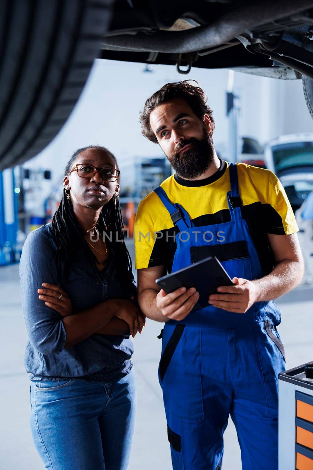 Proficient professional helping customer with car checkup in auto repair shop. Garage employee looking over automobile parts with woman, repairing her busted vehicle wheels during inspection