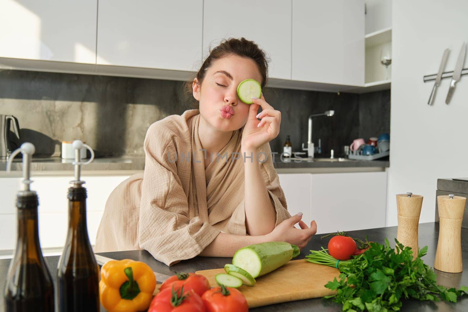 Portrait of cute young woman in bathrobe, cooking meal, standing near chopping board with vegetables, holding zucchini, making salad or vegetarian dinner.