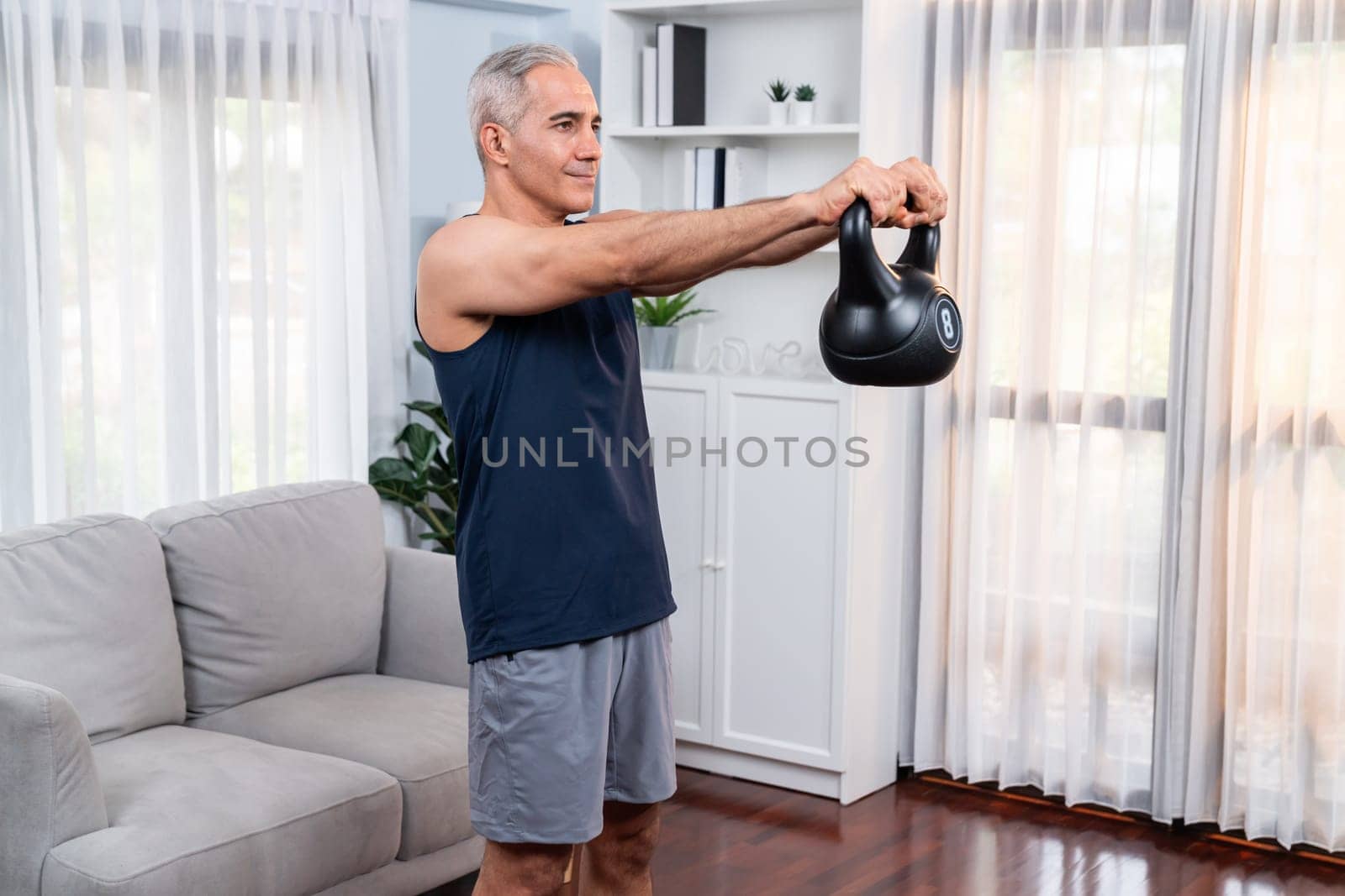 Athletic and sporty senior man engaging in body workout routine with lifting kettlebell at home as concept of healthy fit body with body weight lifestyle after retirement. Clout