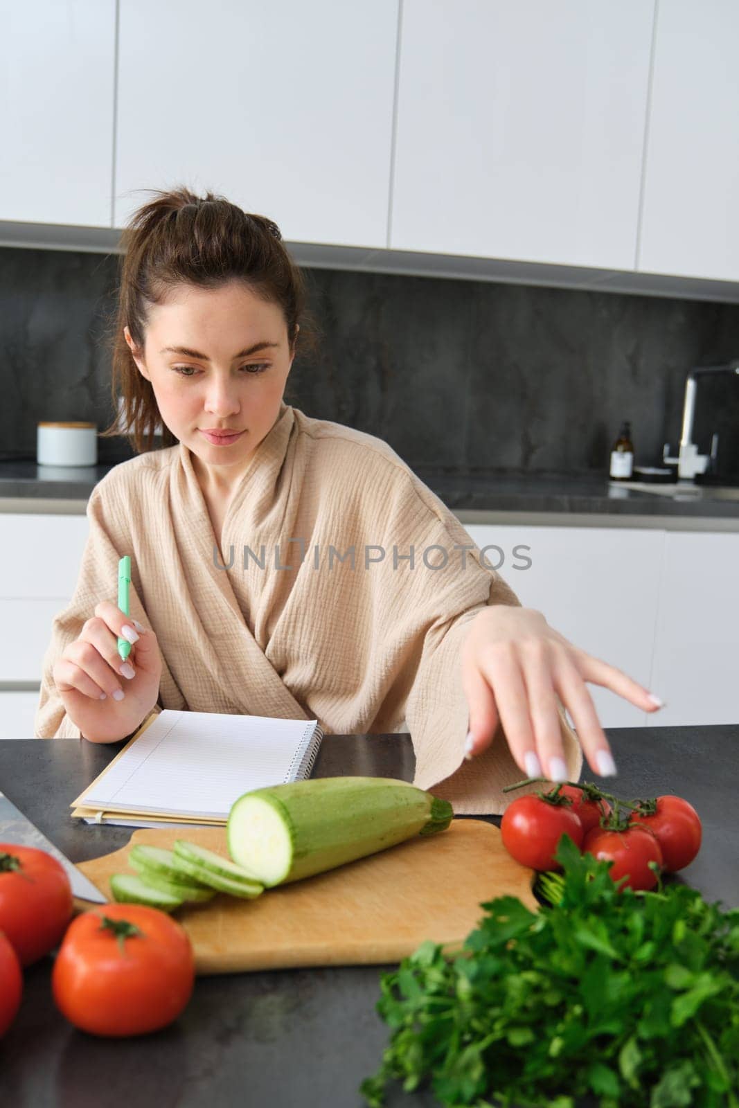Portrait of woman writing down list of groceries, making notes in recipe, sitting in kitchen near vegetables, preparing dinner menu by Benzoix