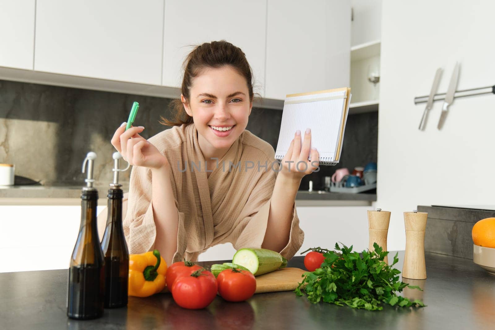 Smiling young woman in bathrobe, cooking in the kitchen, standing near vegetables with notebook, writing down recipe, checking grocery list, cooking fresh and healthy meal by Benzoix