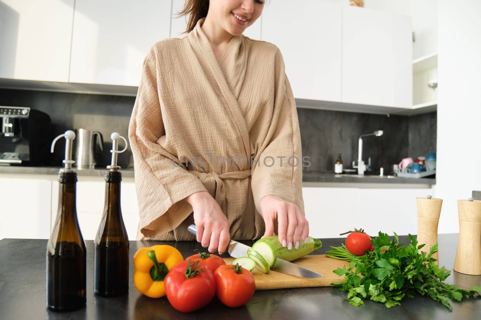 Healthy lifestyle. Young woman in bathrobe preparing food, chopping vegetables, cooking dinner on kitchen counter, standing over white background.