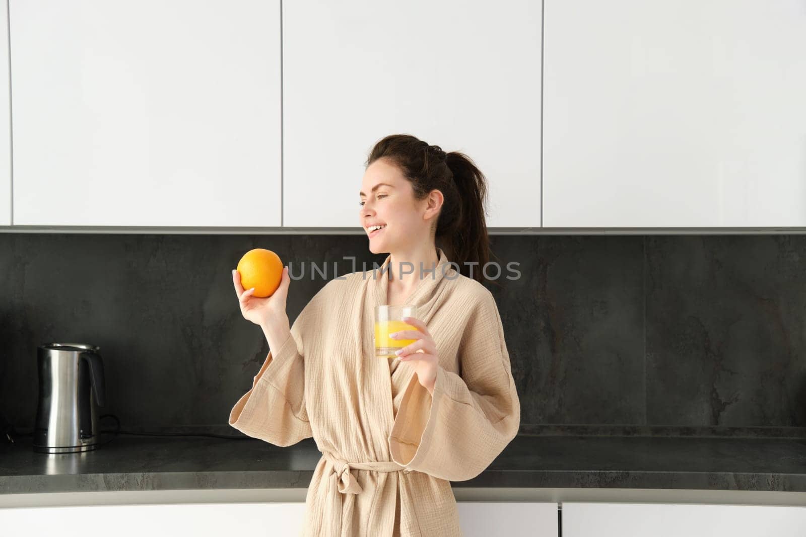 Portrait of attractive young woman, posing with an orange, drinking fresh juice at home in kitchen, starting her morning with healthy drink.