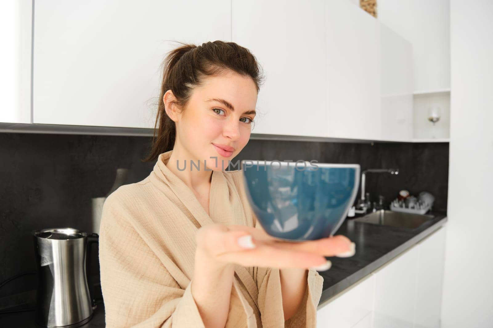 Portrait of beautiful, smiling young woman offering you morning cup of coffee, extending her hand with mug to you, standing in bathrobe in the kitchen.
