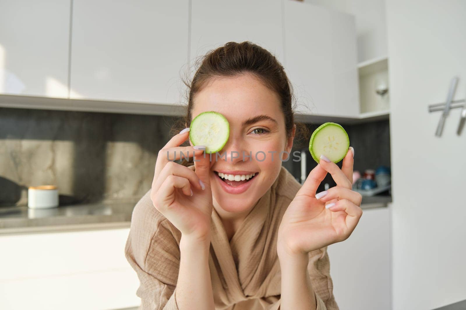 Funny, happy girl eating healthy, holding zucchini, chopping vegetables for healthy meal in the kitchen, posing in bathrobe.