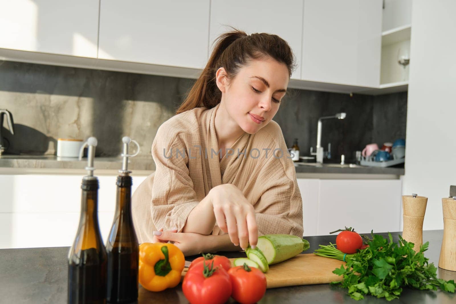 Portrait of cute young woman in bathrobe, cooking meal, standing near chopping board with vegetables, holding zucchini, making salad or vegetarian dinner by Benzoix