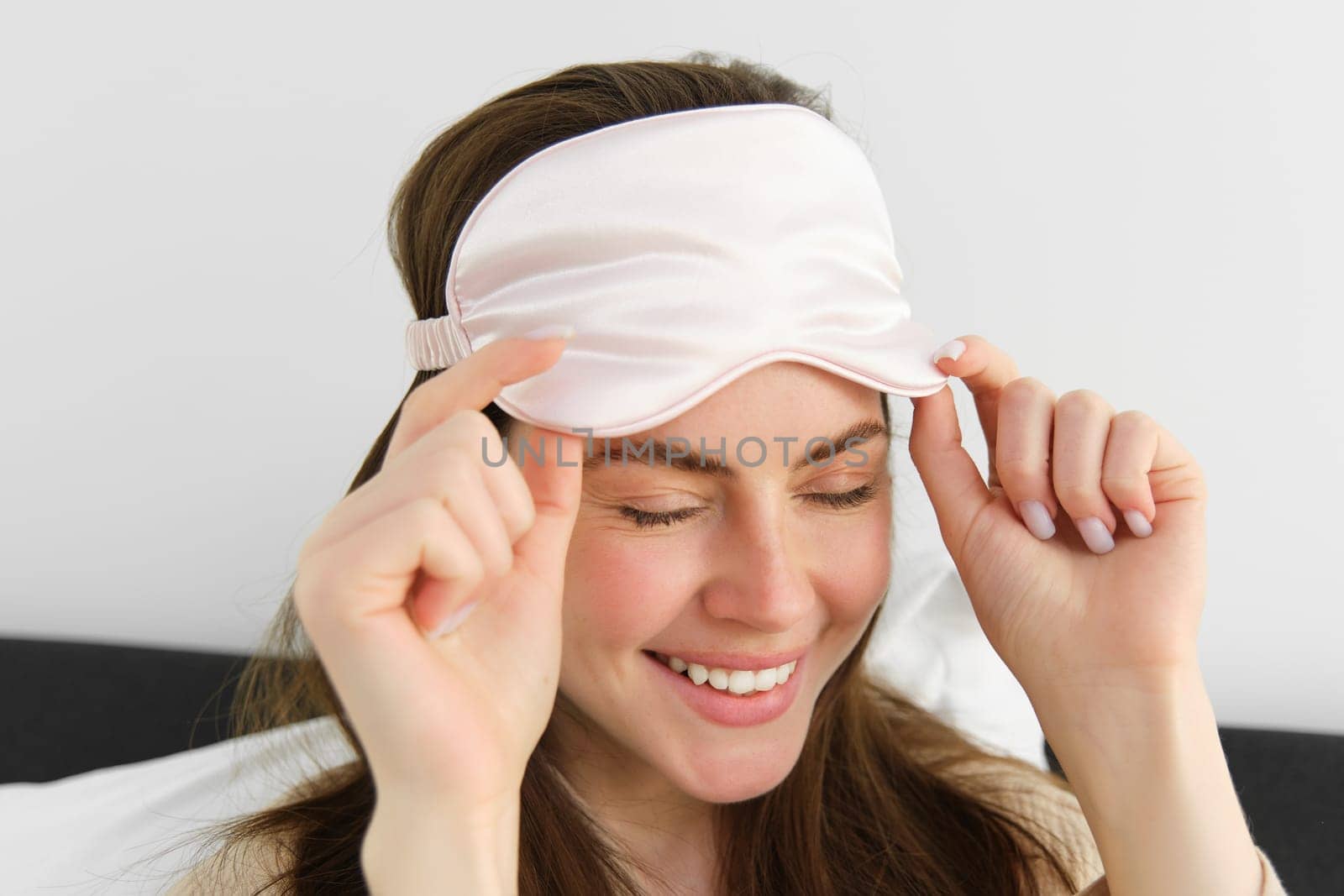 Close up portrait of gorgeous smiling woman with sleeping mask on forehead, waking up in morning, relaxing in bed, laughing and looking happy, resting on vacation in hotel room.