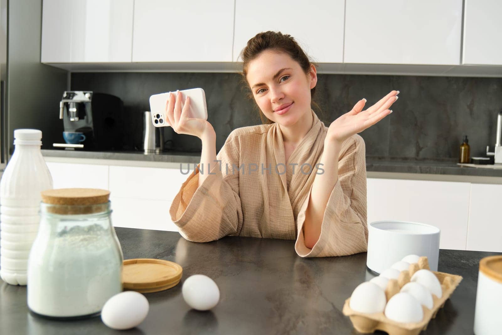 Attractive young cheerful girl baking at the kitchen, making dough, holding recipe book, having ideas by Benzoix