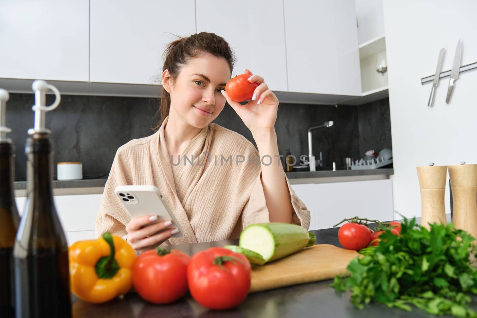 Image of young beautiful woman, holding tomato, sitting in kitchen with smartphone, chopping board and vegetables on counter, cooking food, order groceries for her recipe, using mobile phone app by Benzoix