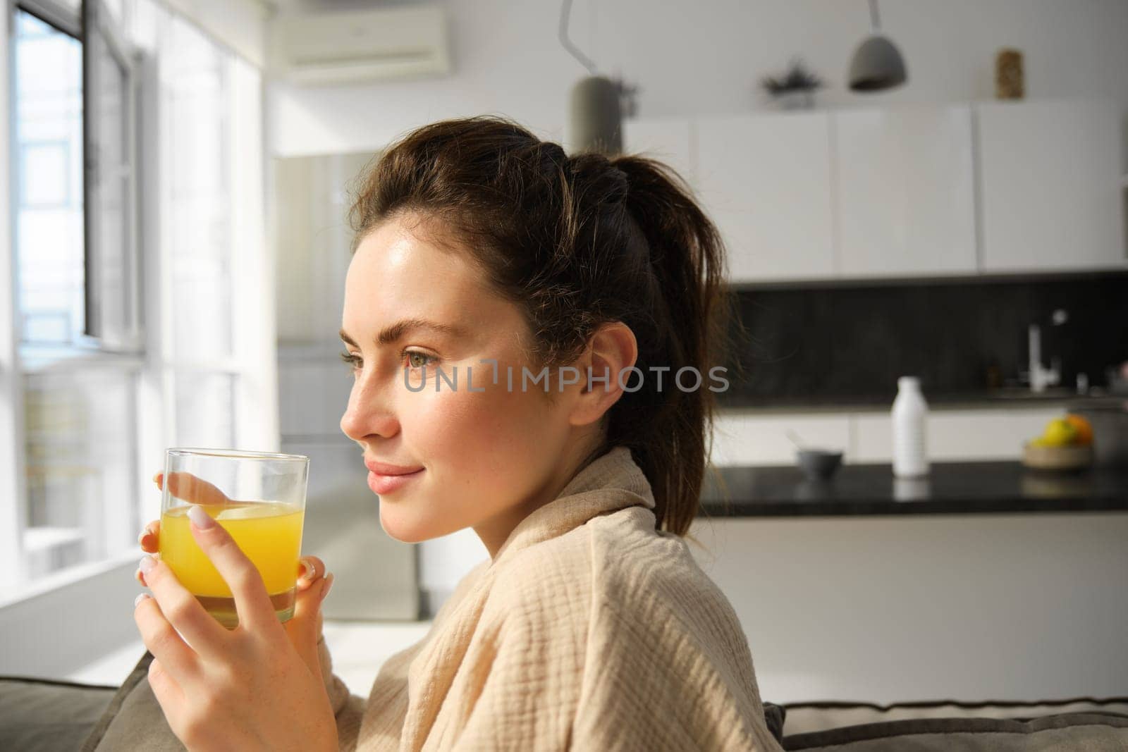 Portrait of gorgeous, modern young woman wakes up in morning, has her fresh glass of orange juice, looks outside window, relaxing in bathrobe at home.