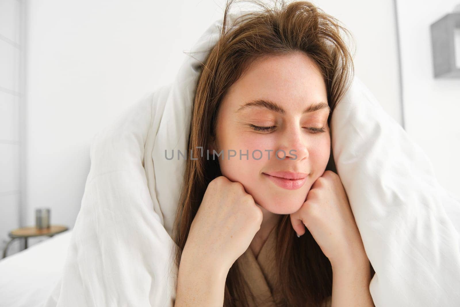 Cheerful woman feeling comfortable in bed, lying in bedroom covered in white sheets, smiling pleased, has messy hair in morning, waking up and looking happy by Benzoix