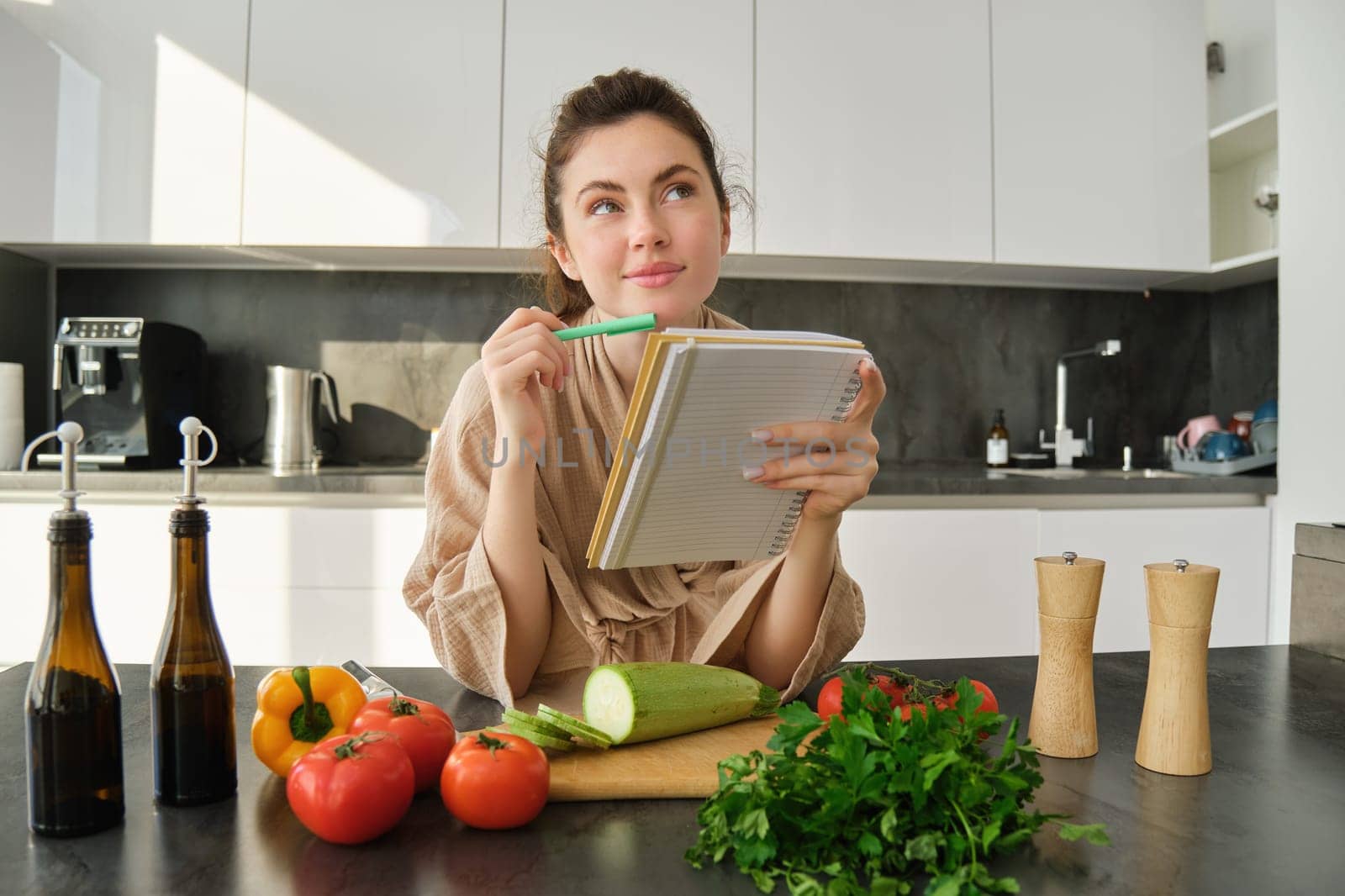 Portrait of woman checking recipe notes in notebook, standing in kitchen with vegetables, cooking food, preparing delicious salad from tomatoes, parsley and olive oil by Benzoix