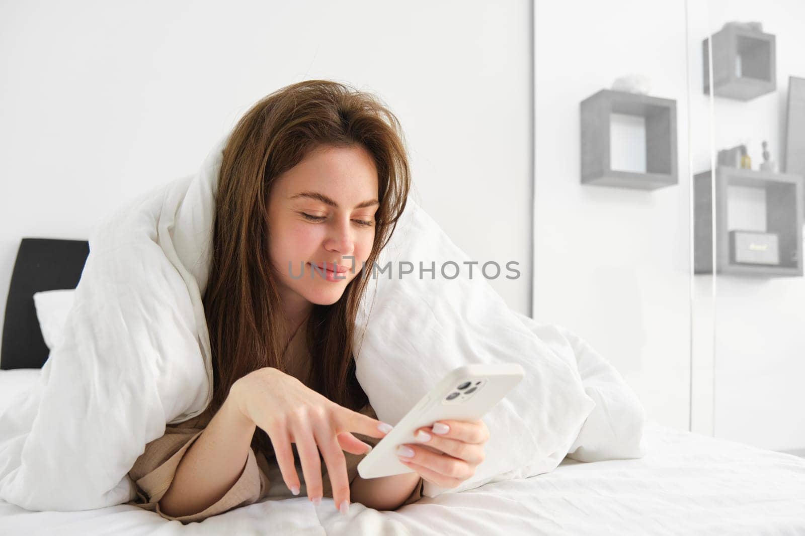 Woman in morning lying in bed covered in blanket, looking at smartphone, using mobile phone app.