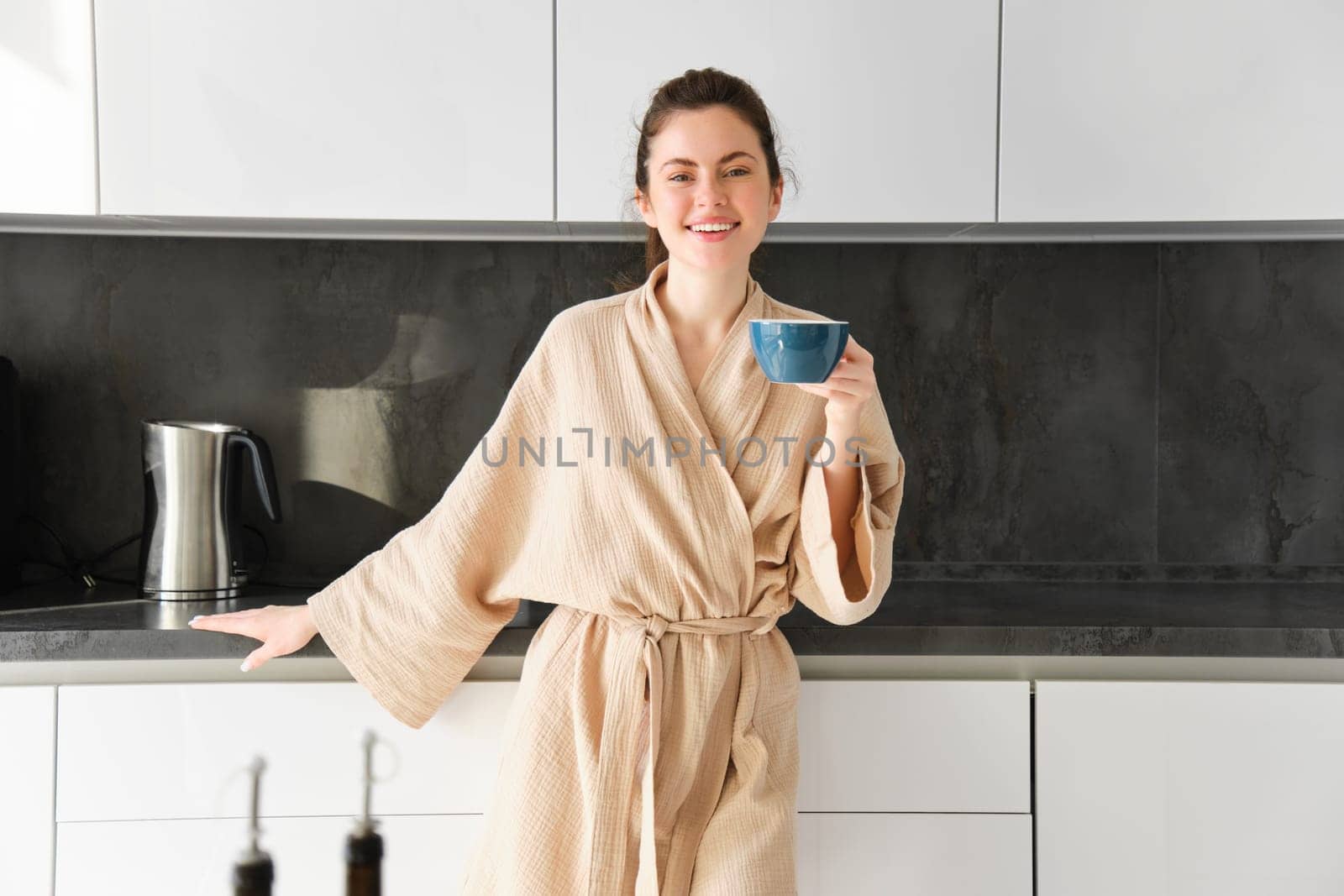 Portrait of beautiful housewife, young woman in bathrobe, holding cup of coffee, drinking tea in the kitchen, enjoying morning.