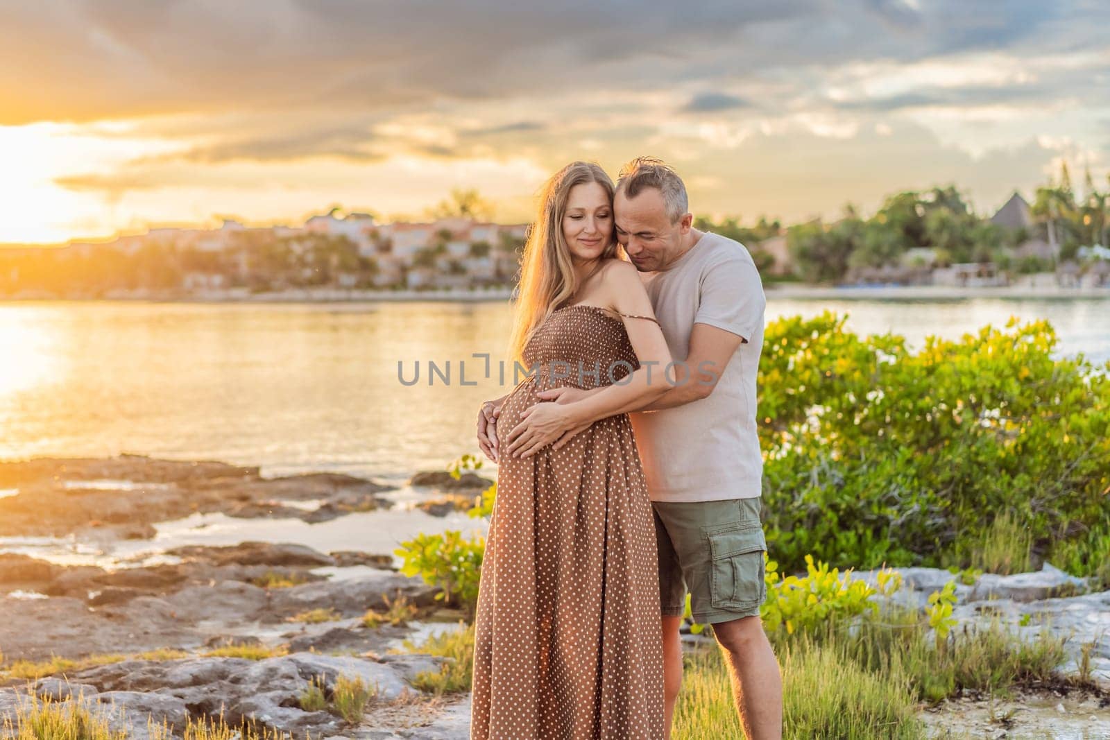 A happy, mature couple over 40, enjoying a leisurely walk on the waterfront On the Sunset, their joy evident as they embrace the journey of pregnancy later in life.