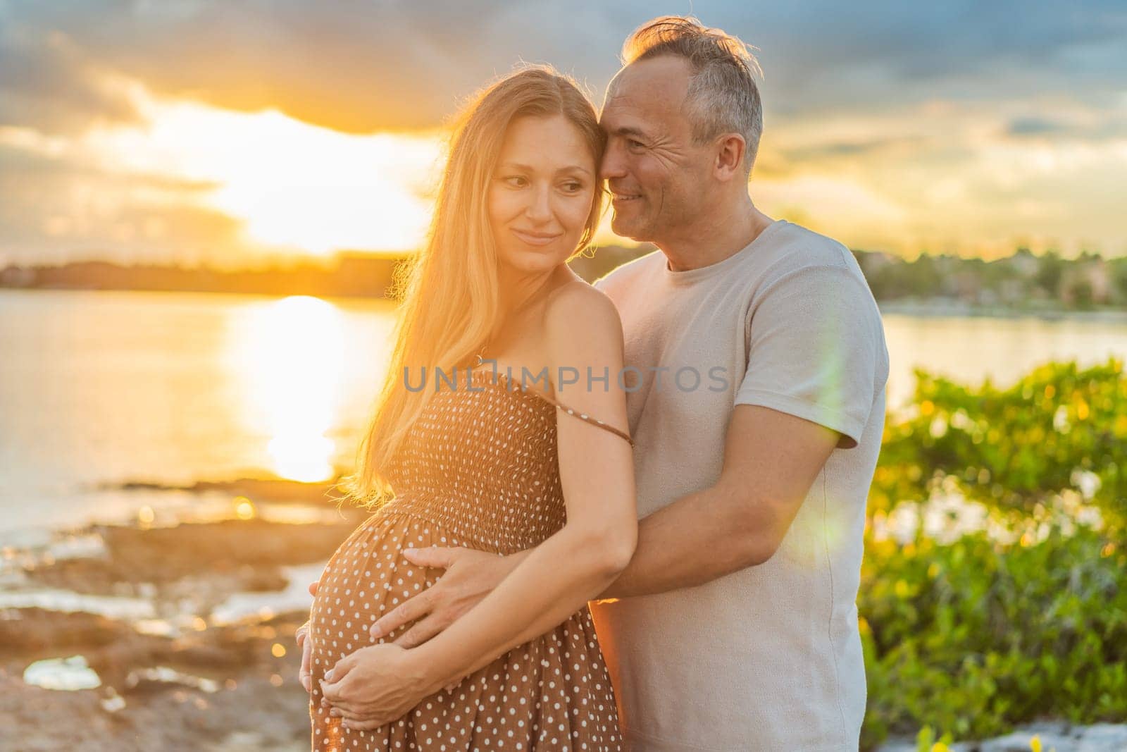 A happy, mature couple over 40, enjoying a leisurely walk on the waterfront On the Sunset, their joy evident as they embrace the journey of pregnancy later in life.