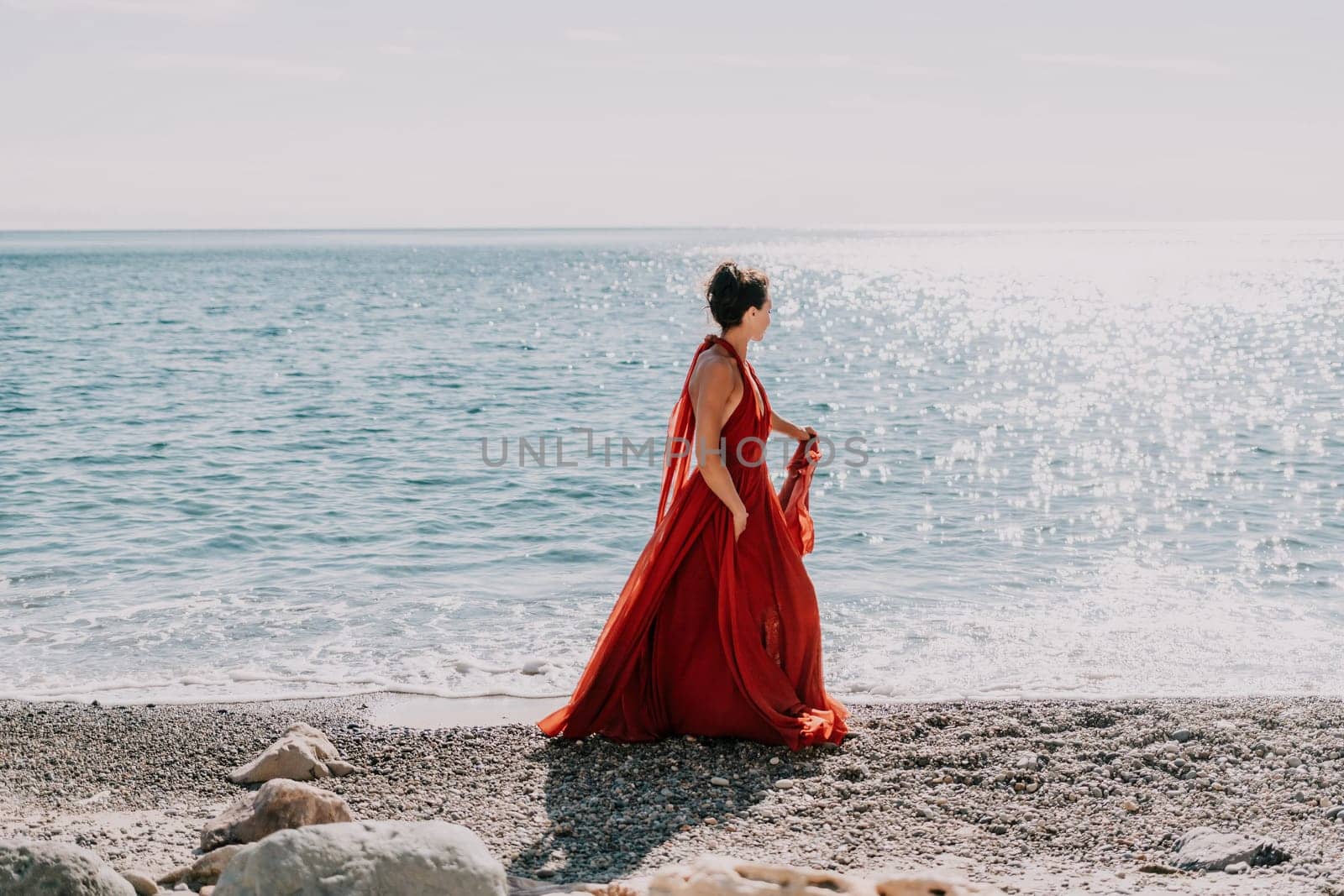 Side view a Young beautiful sensual woman in a red long dress posing on a rock high above the sea during sunrise. Girl on the nature on blue sky background. Fashion photo.