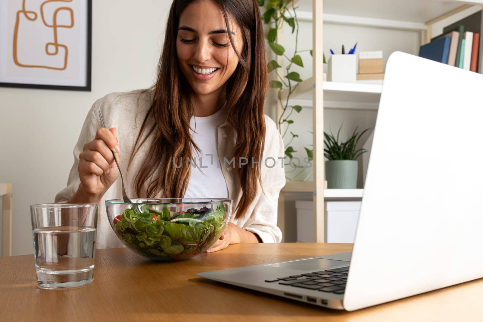 Caucasian woman eating healthy salad for lunch while working with laptop at home office. by Hoverstock