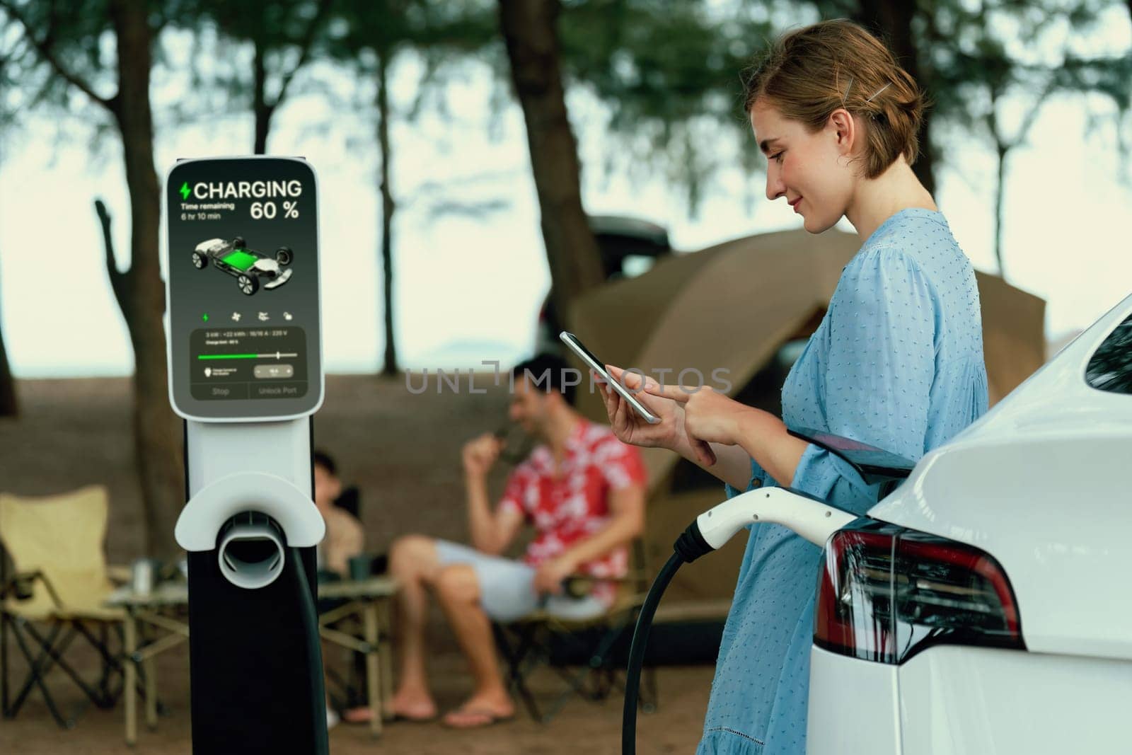 Outdoor adventure and family vacation camping at sea travel by eco friendly car. Woman or mother check car's battery with smartphone while charging EV car frin charging station in campsite. Perpetual