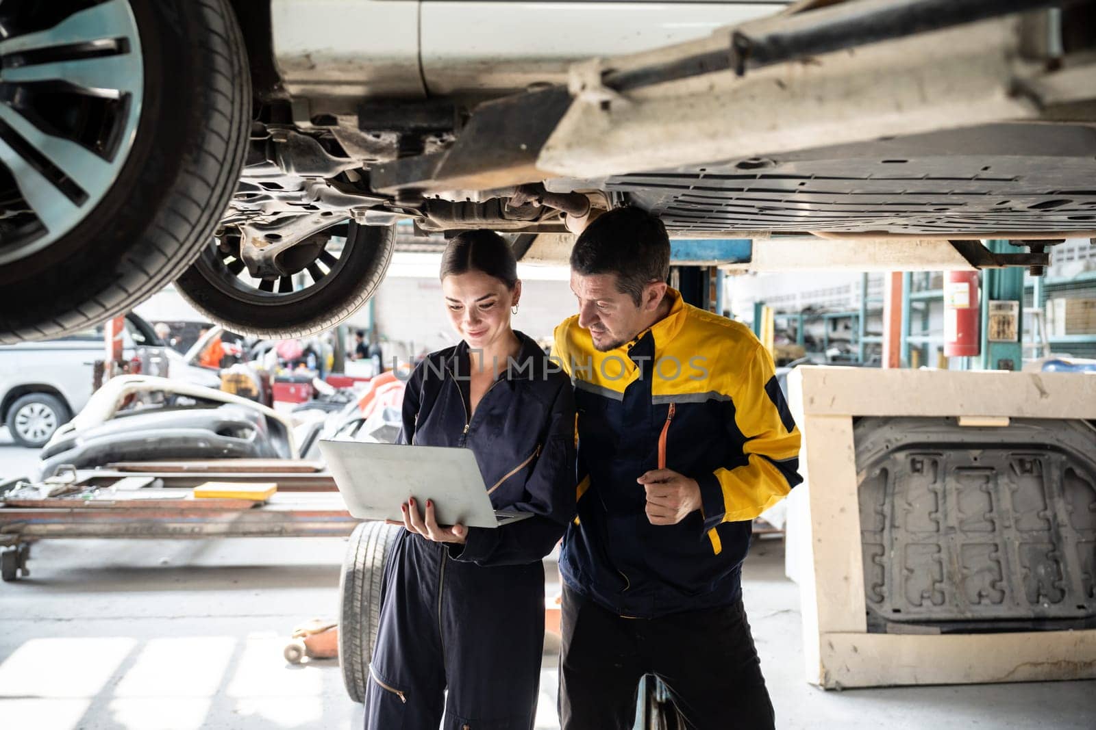 Two vehicle mechanic working together underneath lifted car, conduct car inspection with laptop. Automotive service technician in uniform carefully make diagnostic troubleshooting. Oxus