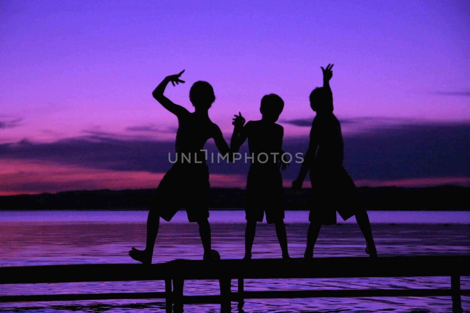 Three silhouettes of boys on a dock against a sunset