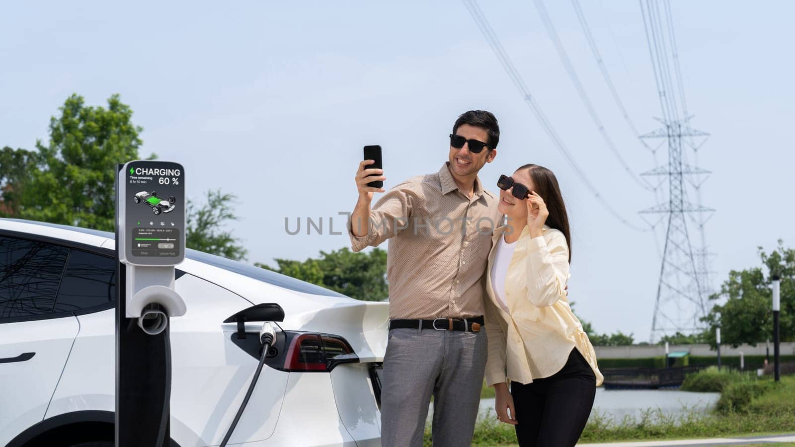 Couple pay for electricity with smartphone while recharge EV car battery at charging station connected to power grid tower electrical as electrical industry for eco friendly car utilization.Expedient