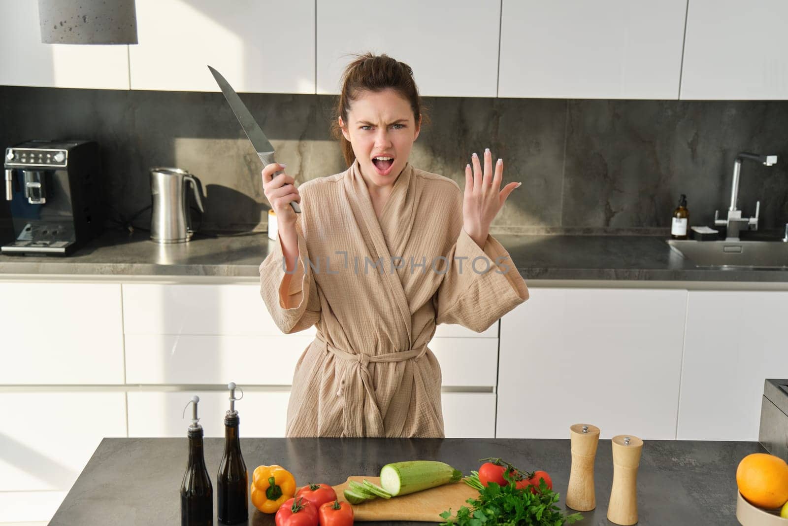 Portrait of annoyed woman with knife, angry while cooking in the kitchen, frustrated while doing house chores and preparing food for family, standing in bathrobe.