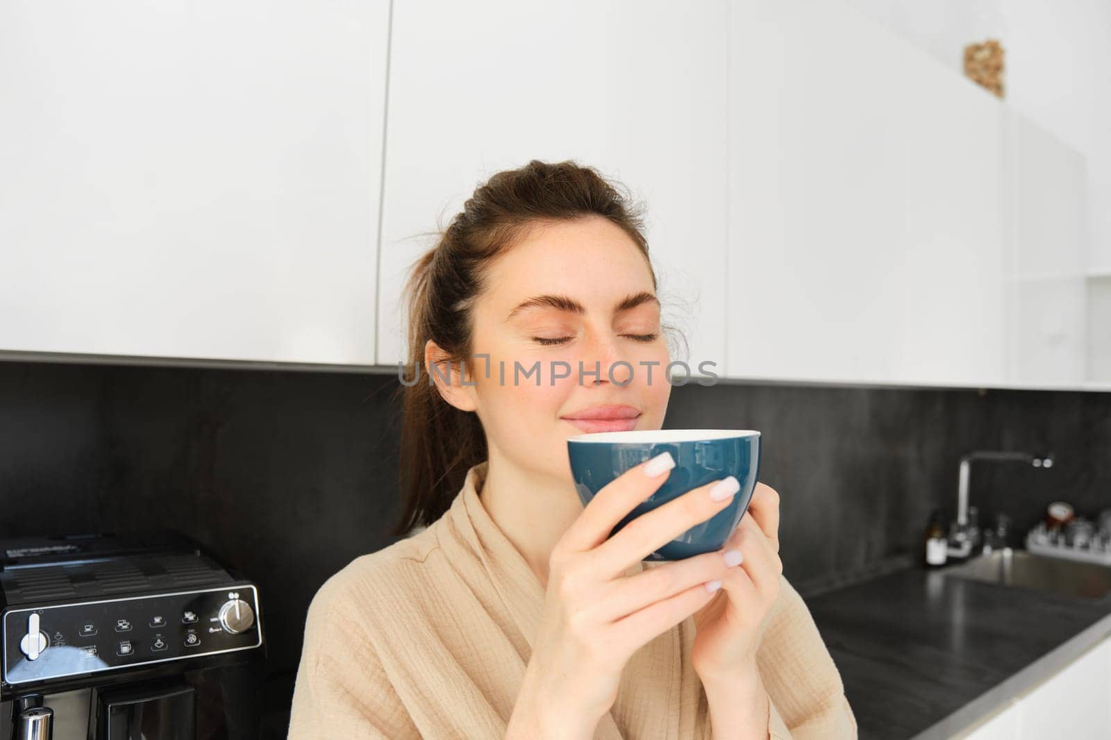 Portrait of beautiful young woman, drinking coffee, freshly made cappuccino, smiling pleased, enjoying her morning, standing in the kitchen.