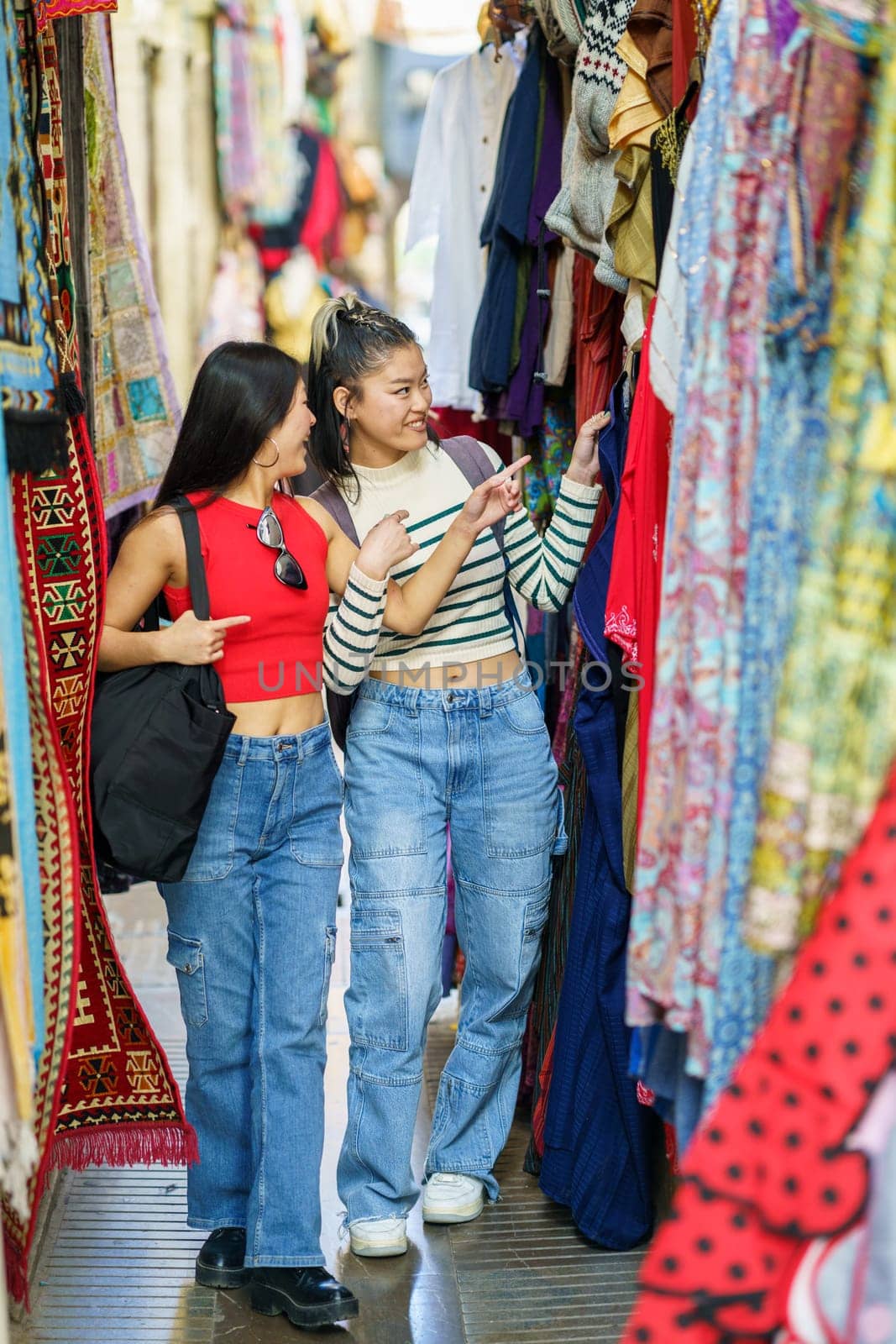 Smiling girls friends in casual clothes walking along colorful clothing store on street market in city during summer vacation
