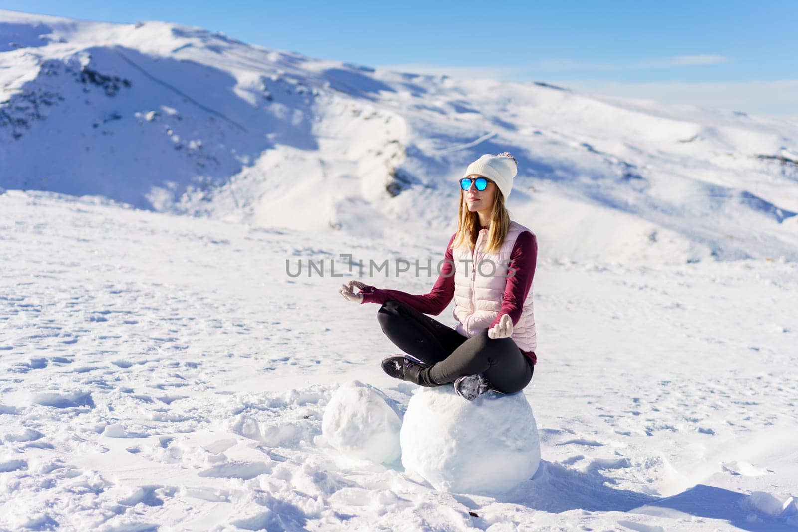 Full body of young female traveler in casual clothes and sunglasses sitting on snow in lotus pose and looking away on sunny winter day