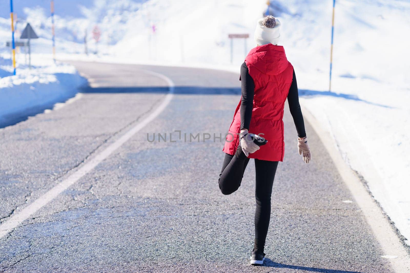 Back view of anonymous athletic lady in warm clothes stretching legs while standing on asphalt road in snowy mountains