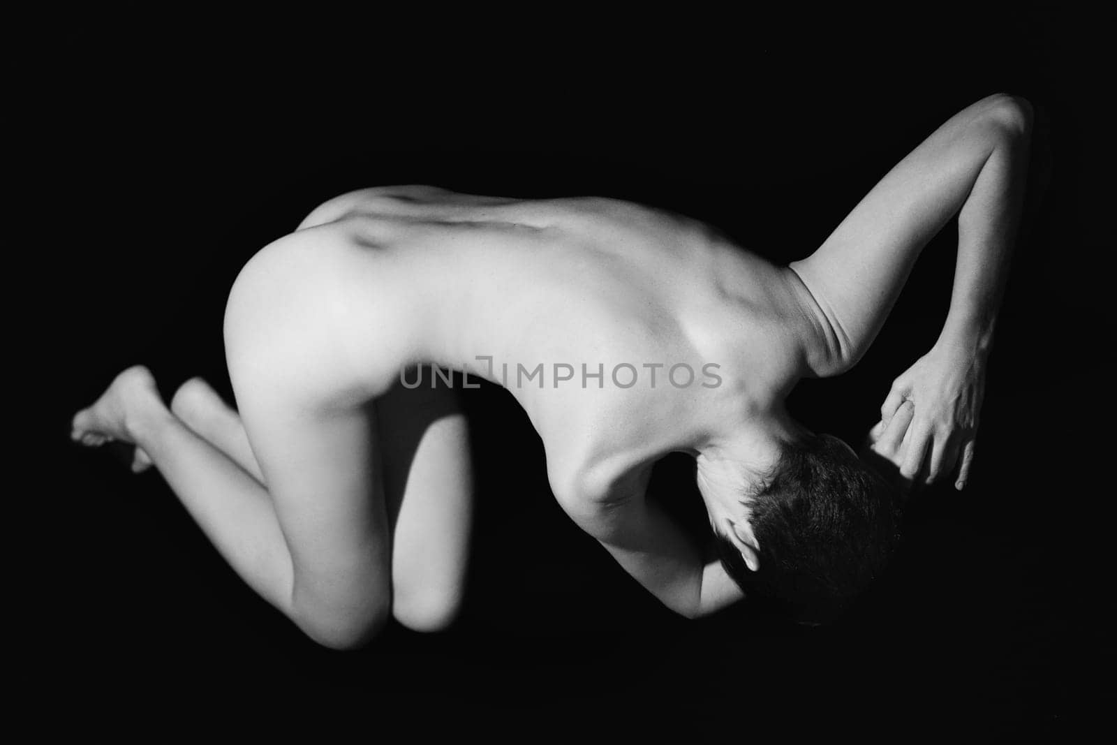 Full body of anonymous fit naked woman with short dark hair lying on floor against black background in studio. Black and white photograph