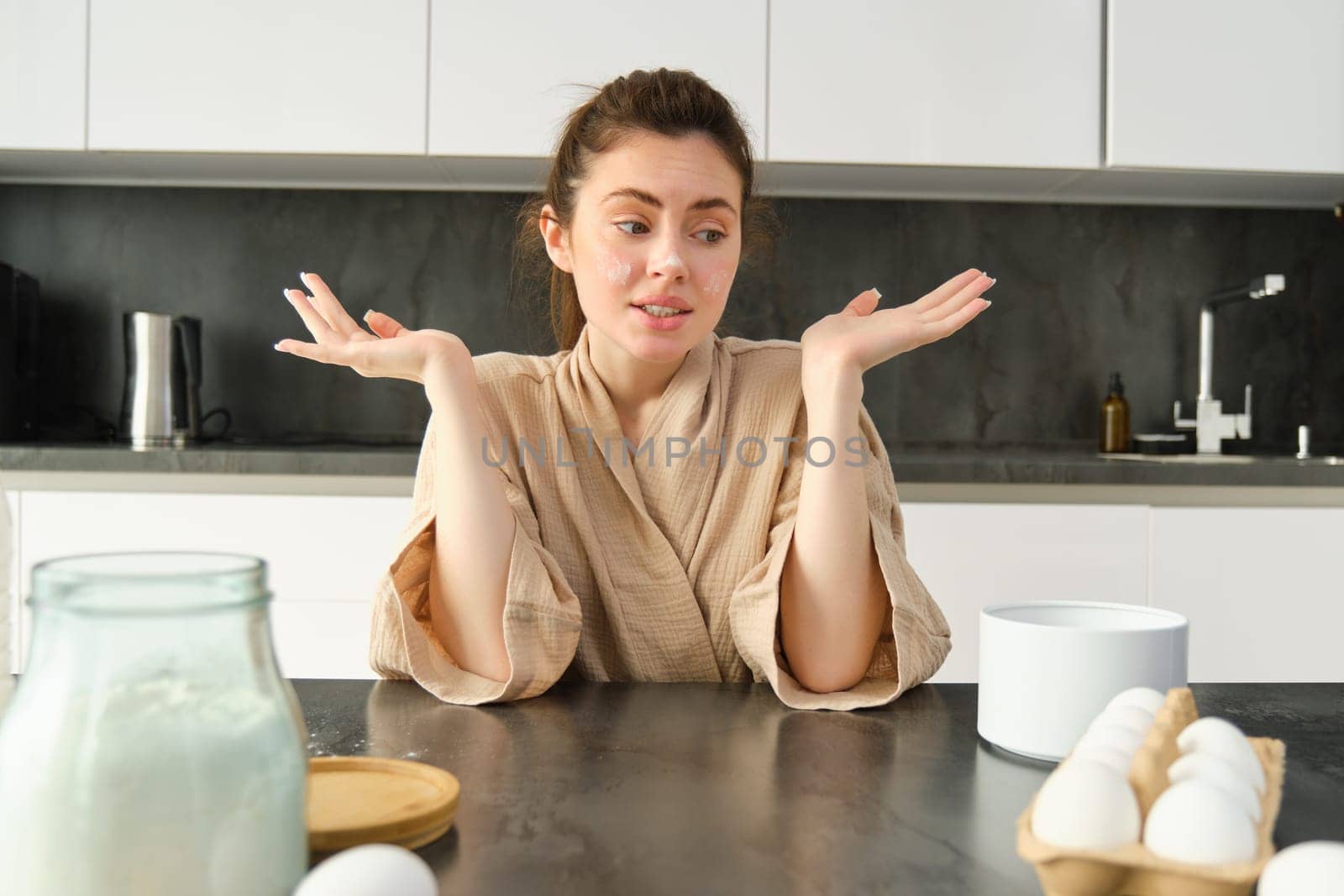 Attractive young cheerful girl baking at the kitchen, making dough, holding recipe book, having ideas.