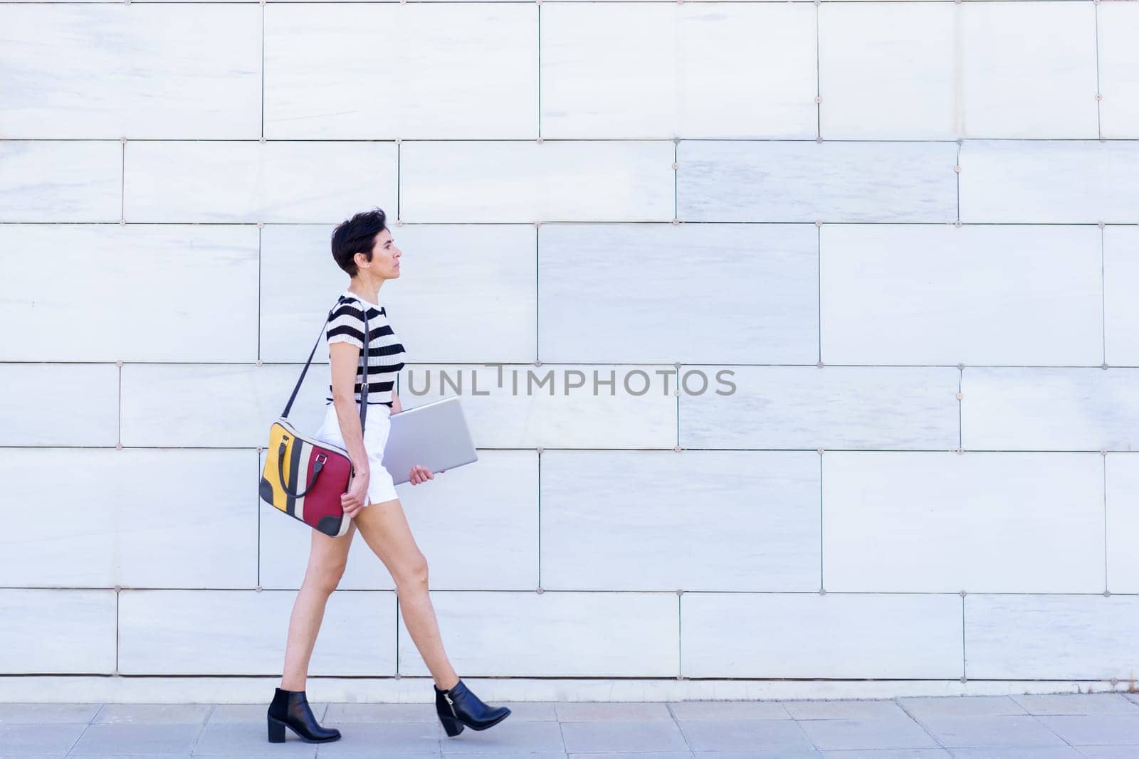 Full body side view of pensive young female in striped t shirt and shorts with bag and laptop walking on city street in daytime