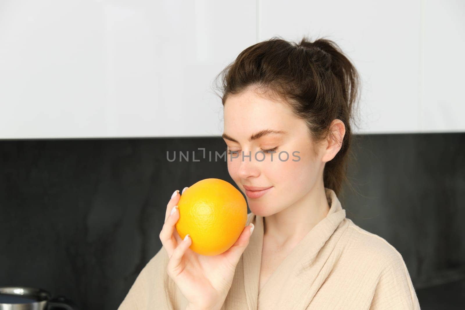 Close up portrait of young brunette, smells fresh orange, drinking juice from glass, enjoying healthy start of morning, wearing bathrobe by Benzoix