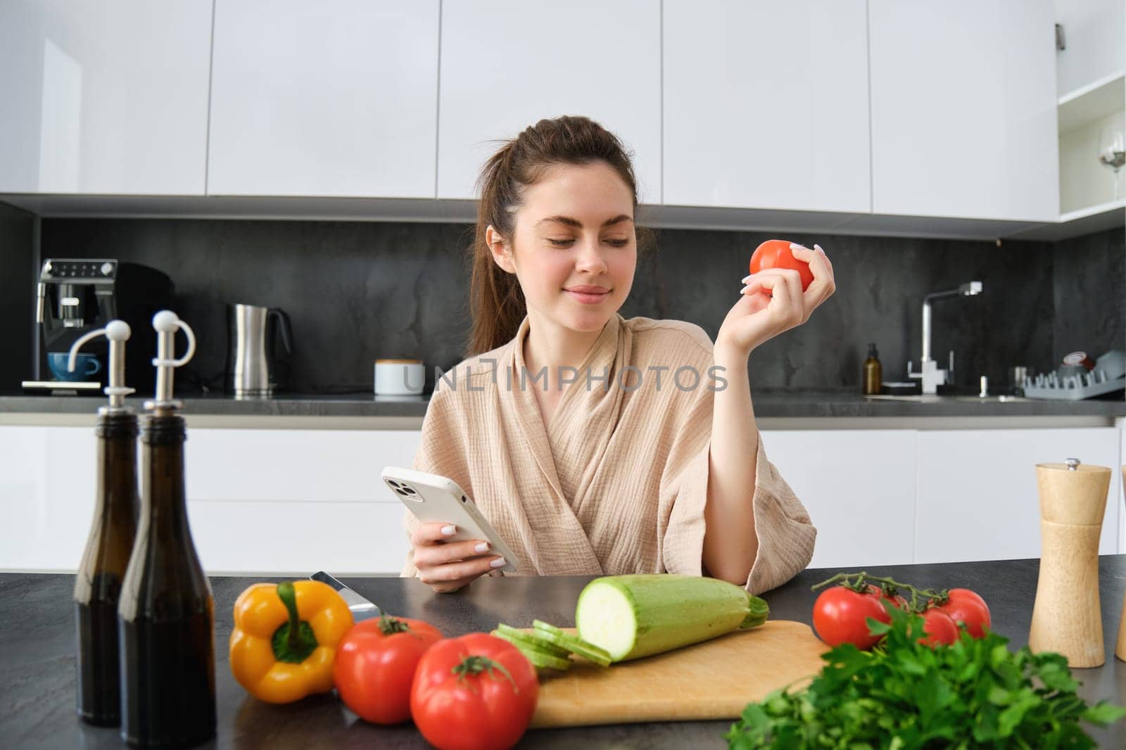 Young woman orders groceries on mobile app. Girl in bathrobe sits in the kitchen with vegetables, looking for recipe to cook dinner, using smartphone application.