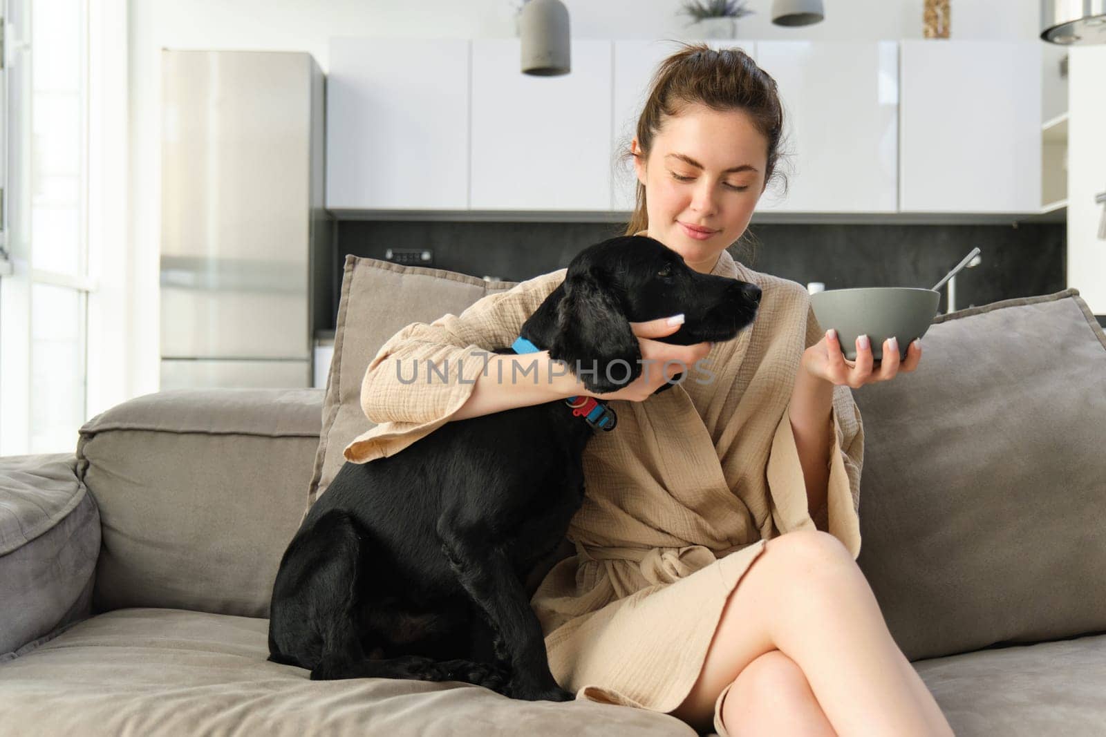 Portrait of cute dog asking for girls breakfast, looking at bowl of cereals. Young woman doggo owner eating on sofa, wearing bathrobe.