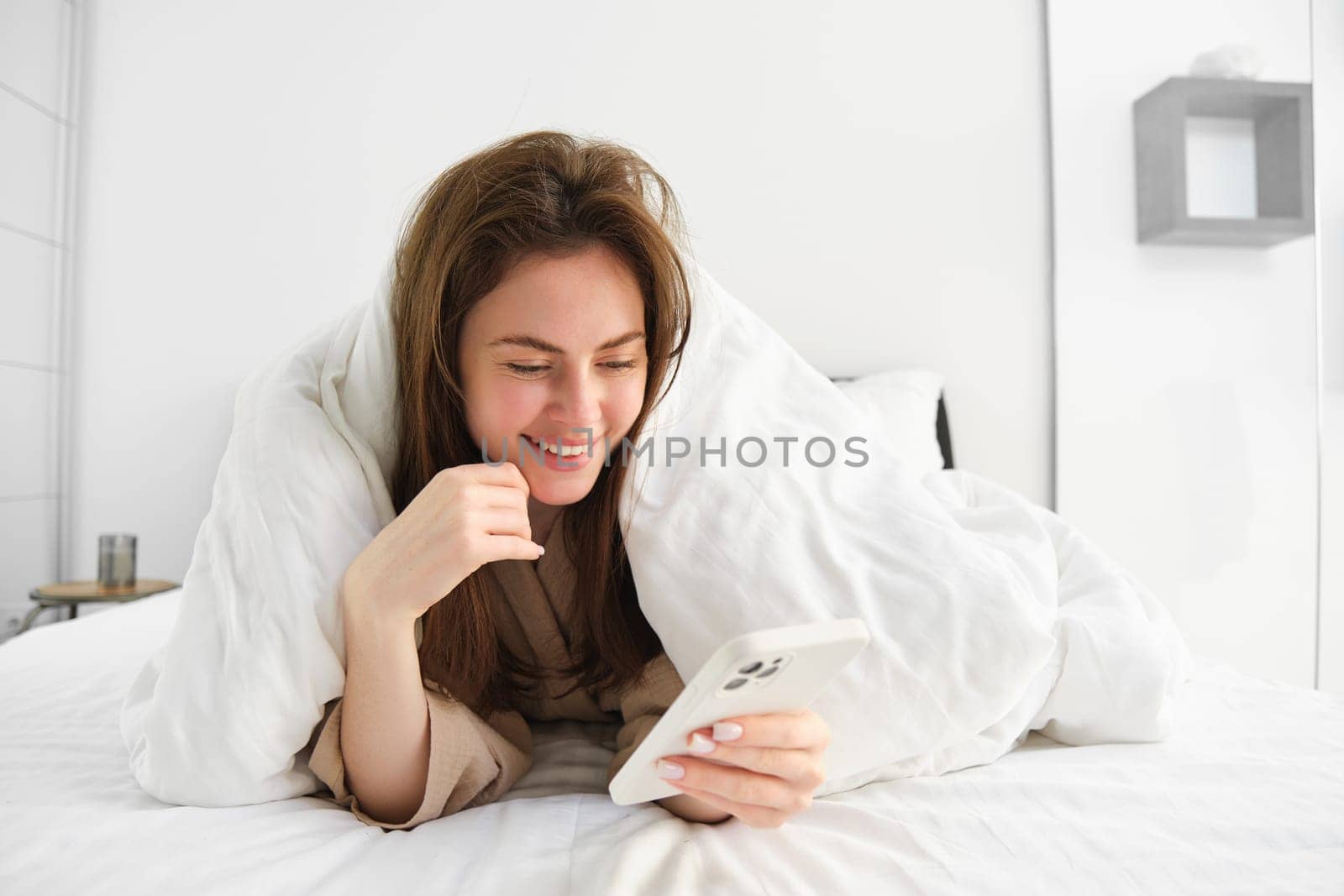 Happy woman in bed, looking at smartphone, online shopping while relaxing in her bedroom under blankets.