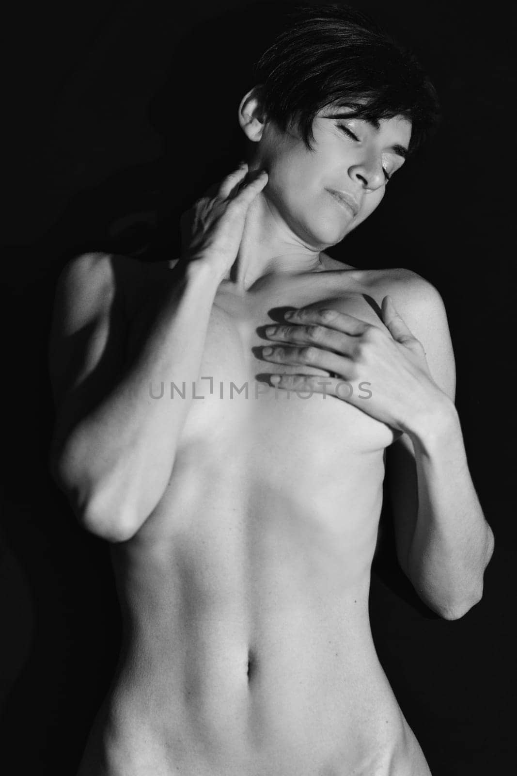 Black and white of sensual adult female with short dark hair covering naked breast and touching neck with closed eyes in studio