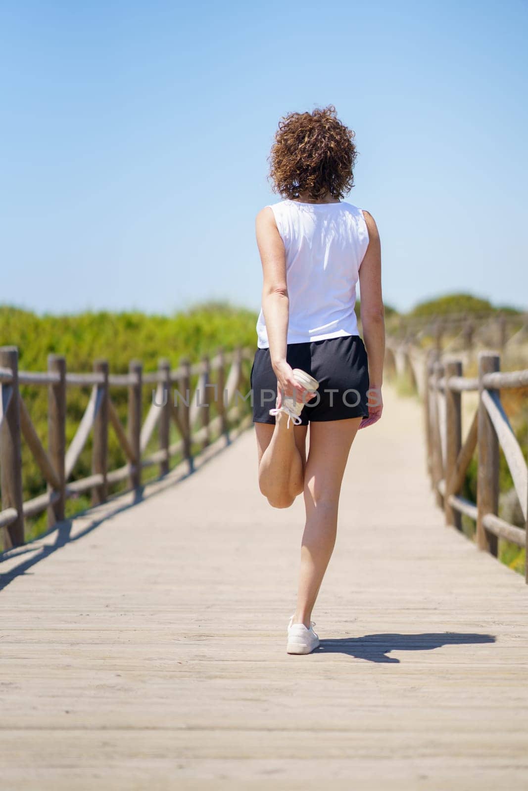 Full body back view of anonymous female athlete in sportswear stretching leg on wooden walkway against blue sky on sunny summer day