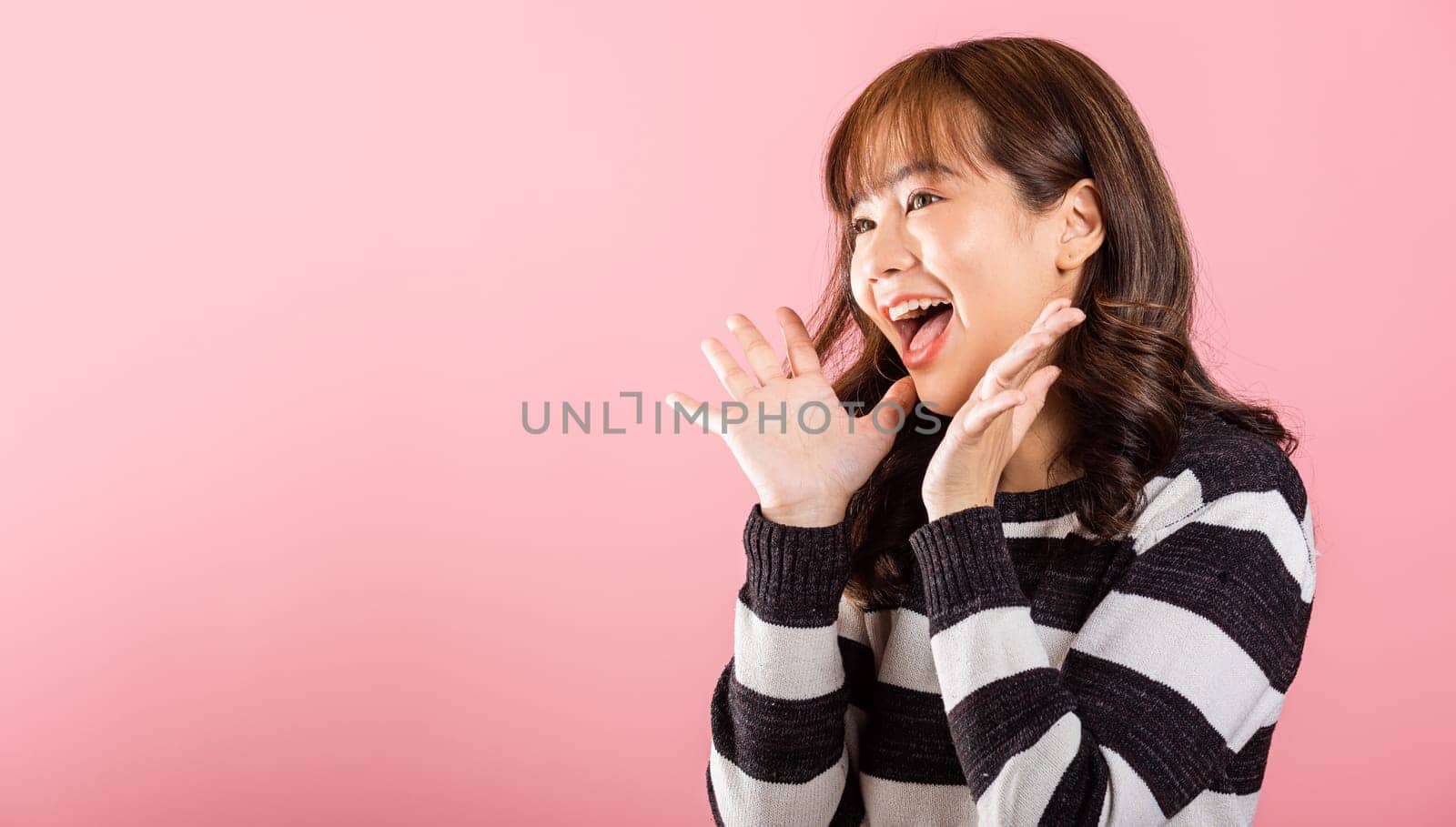 An Asian woman smiles with joy, making a victorious gesture by raising her fists. Studio shot on a pink background, radiating success and excitement.