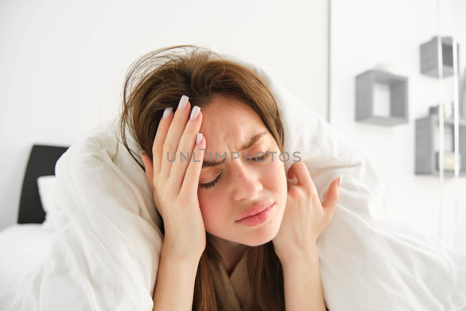 Portrait of woman with headache, lying in bed and touching head with frustrated, frowning face, does not feel well, stays in bedroom as having migraine in morning by Benzoix