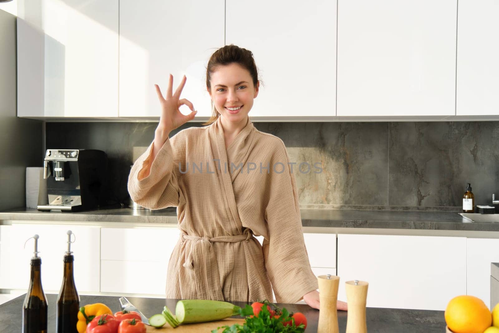 Portrait of smiling brunette woman cooking healthy dinner, showing okay sign, wife standing in bathrobe in kitchen, preparing food, chopping vegetables on board by Benzoix