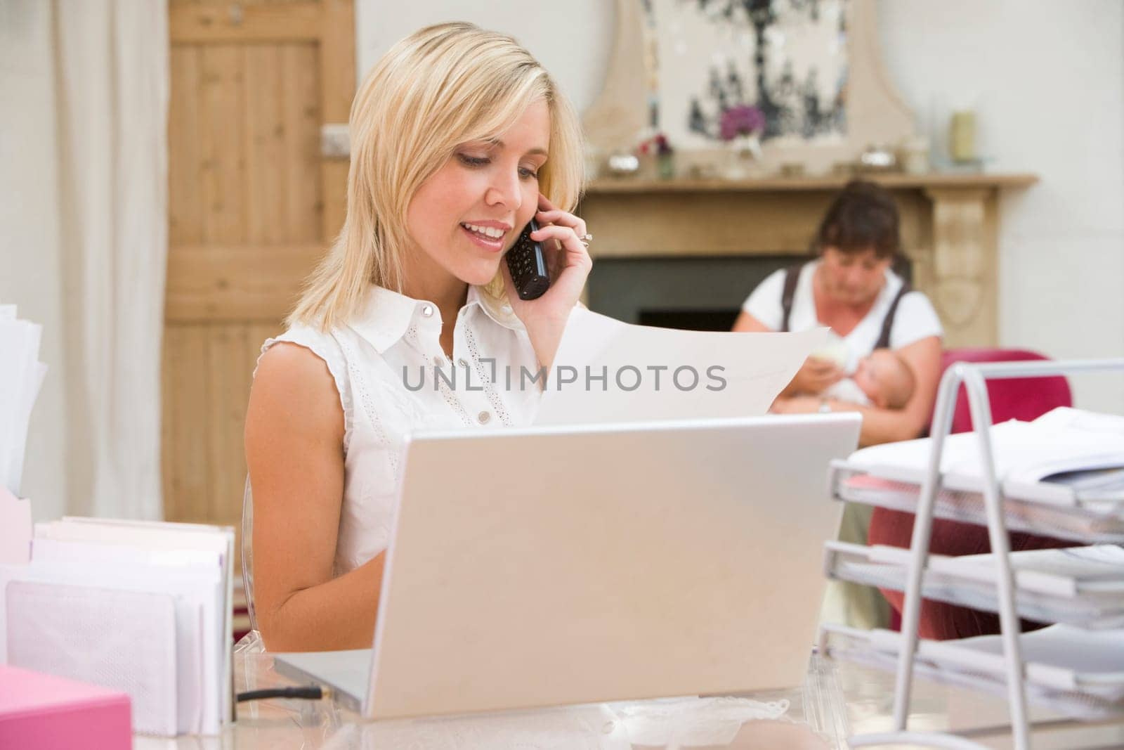 Woman in home office with laptop and telephone with mother and baby in backgroun