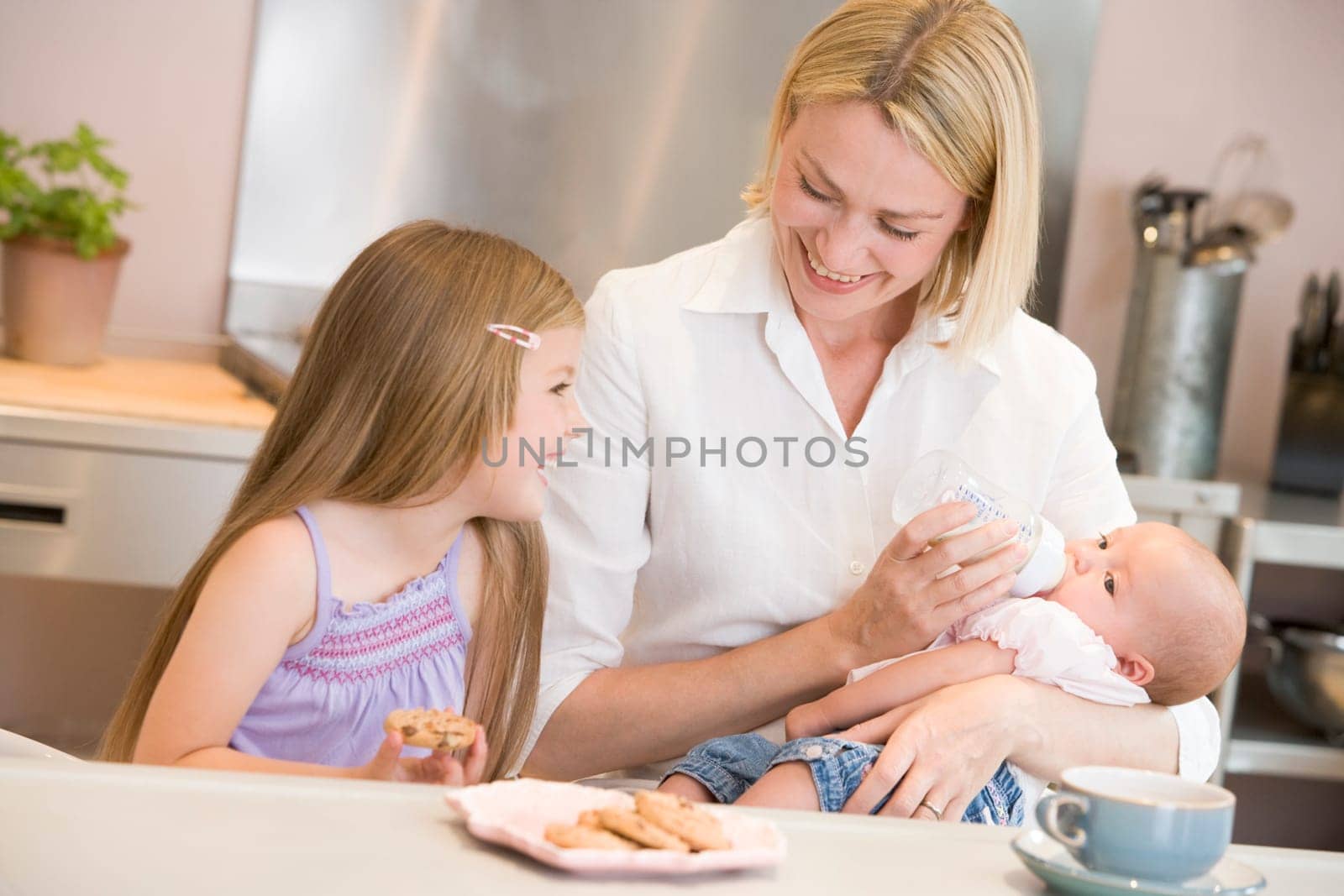 Mother feeding baby in kitchen with daughter eating cookies and smiling